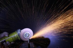a close up of a hand holding an angle grinder to polish a pice of metal.sparkls fly from the point of contact.the worker is holding the grinder with one hand photo