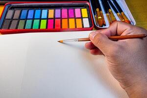 Painting in blank drawing book with brush and watercolor color palette. Paint accessories and watercolor And Blank Book on Wooden Table. Flat lay, top view. Place for text. creative concept photo