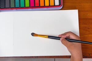 Child painting in blank drawing book with brush and watercolor color palette. Paint and watercolor accessories and Blank Books on Wooden Table. Flat lay, top view. Place for text photo