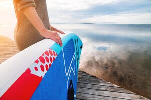 Close-up of a woman's hands lying on the side of a paddleboard. A woman stands on the shore with a wooden pier. Preparing for a walk on the water on a paddleboard photo