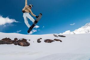 snowboarder jumps into the air against a background of blue sky and mountains. The guy grabs the edge of the board with his front hand while jumping, performing a trick photo
