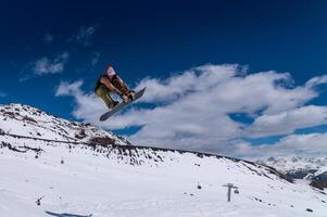 snowboarder jumps from a big air with a board grab, a nose or tail trick in rotation, against the backdrop of snow-capped mountains and the bright sun photo