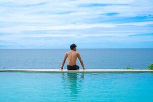 A man relaxing in a swimming pool at a luxury resort with blue sky and blue sea background. Young man enjoying beautiful sunset in luxury hotel near swimming pool. Calmness and serenity. photo