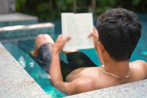 Asian man laying in the swimming pool reading a book in the summer on a sunny day. Relaxing in a backyard swimming pool for relax in summer vacation day. photo