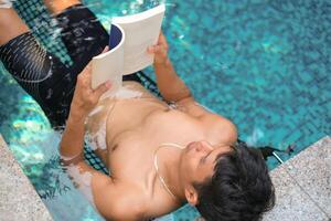 Asian man laying in the swimming pool reading a book in the summer on a sunny day. Relaxing in a backyard swimming pool for relax in summer vacation day. photo