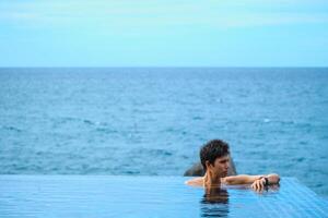 A man relaxing in a swimming pool at a luxury resort with blue sky and blue sea background. Young man enjoying beautiful sunset in luxury hotel near swimming pool. Calmness and serenity. photo
