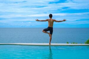 Man doing yoga and meditating in tree position near swimming pool on the tropical beach in Samui, Thailand. Healthy lifestyle active balanced practicing meditate and energy, yoga in morning. photo