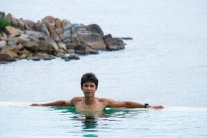 A man relaxing in a swimming pool at a luxury resort with blue sky and blue sea background. Young man enjoying beautiful sunset in luxury hotel near swimming pool. Calmness and serenity. photo