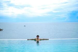 A man relaxing in a swimming pool at a luxury resort with blue sky and blue sea background. Young man enjoying beautiful sunset in luxury hotel near swimming pool. Calmness and serenity. photo