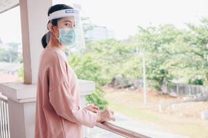 Asian woman wearing mask and face shield before going outside her room for prevent and protection covid-19 virus disease. Conceptual of new normal lifestyle after Covid-19 pandemic outbreak. photo