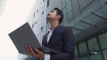 A man in a suit holding a laptop in front of a building video