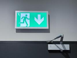 A green emergency exit sign with a figure running and a large arrow pointing down, mounted above a door closer mechanism on a gray wall photo