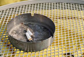Two cigarettes on metal iron ashtray with ashes isolated on horizontal ratio yellow fabric and white metal table surface background. photo