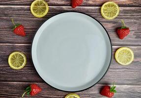 Empty white round plate with plastic lemon slices and strawberry fruits around the plate isolated on horizontal wooden surface background. photo