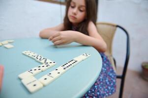 Young girl playing dominoes at a table, focused on aligning the pieces. Captures the essence of childhood concentration and leisure in a calm, indoor setting. photo