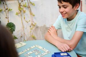 Smiling boy playing dominoes at a table outdoors, enjoying a fun and engaging game with friends or family in a sunny garden setting, showcasing leisure and happiness. photo