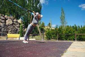 Woman exercising outdoors, practicing resistance training in a park setting, showcasing strength, balance, and determination under a clear blue sky with trees and rock formations. photo