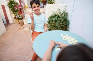 Smiling boy playing dominoes with a friend in a sunny outdoor patio, enjoying a fun and relaxed game, surrounded by plants and cheerful atmosphere photo