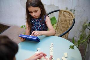 Young girl playing dominoes at a table, concentrating on her next move during a calm game session. Child enjoying a classic board game outdoors in a relaxed setting. photo