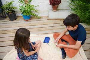 Children playing domino game outdoors on wooden deck, enjoying leisure time and bonding on a warm day with plants surrounding them photo