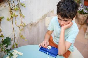 Pensive boy engaged in a thought-provoking game of dominoes at a round table, surrounded by outdoor elements and a hint of greenery, reflecting concentration and contemplation. photo