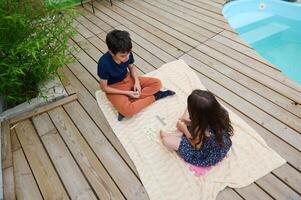 Children enjoying a fun game of word tiles on a cozy blanket by the poolside, creating a perfect summer day scene filled with laughter and creativity. photo