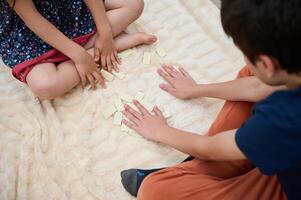Children playing a game with dominoes on a soft beige blanket, focusing on hands and game pieces, creating a cozy and playful atmosphere at home. photo
