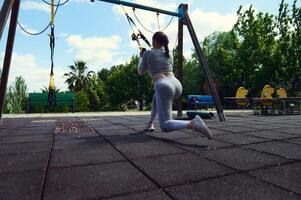 Woman exercising with TRX straps in outdoor park setting, showcasing strength and fitness in a natural environment, with trees and playground equipment in the background. photo
