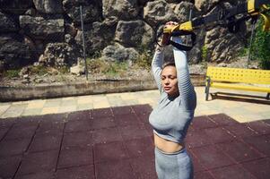Young woman exercising with suspension trainer outdoors in urban park setting on a sunny day photo