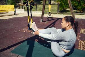 Woman exercising with TRX suspension trainers outdoors on a sunny day, focusing on fitness and core strength with dedication and concentration photo
