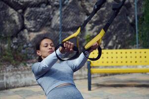Woman exercising with TRX straps in an outdoor setting, focusing on strength and fitness. Yellow bench and rocky background create a dynamic workout environment. photo