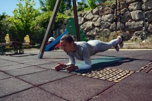 Woman performing a forearm plank exercise on a mat in an outdoor playground setting, exemplifying strength, balance, and fitness routines in a natural environment. photo