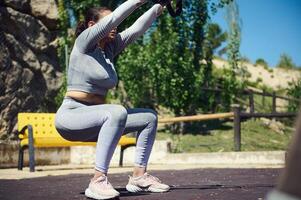 Woman performing outdoor squat exercise with resistance bands in a sunny park setting, focusing on fitness and strength training in a natural environment. photo