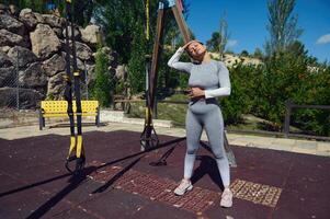 Woman stretches outdoors in fitness gear, enjoying a sunny day workout. TRX bands and serene park setting create perfect fitness atmosphere. photo