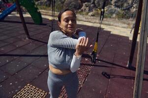 Woman in athletic attire stretching her arm outdoors in a park setting, enjoying a sunny day. Focus on fitness and wellness in an outdoor environment. photo