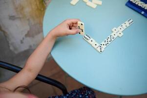 Child playing dominoes at a round blue table outdoors, developing strategic thinking and motor skills while enjoying leisure time in a casual setting on a sunny day. photo