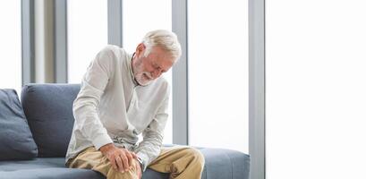 Senior man suffering from knee pain sitting sofa in the living room, Elderly man suffering from knee pain while sitting on the sofa photo