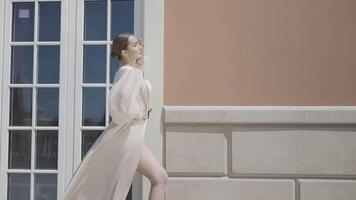 Side view of a gorgeous female model in a silk dress swaying in the wind. Action. Woman standing and posing in front of a building with big window and brown facade. video