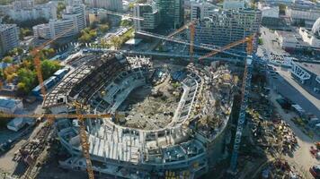 Top view of construction of sports arena with cranes. Stock footage. Construction of sports arena in center of modern city in summer. A lot of construction cranes on site of sports arena in city on video