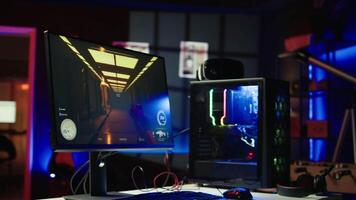 Panning shot of empty neon lit living room with RGB lit computer on table next to headphones and high tech VR headset. FPS videogame left open on pc display and computer peripherals in apartment video