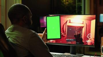 Man watching videos on green screen tablet while playing first person shooter videogame at home. Gamer looking at chroma key device display while enjoying online game on PC