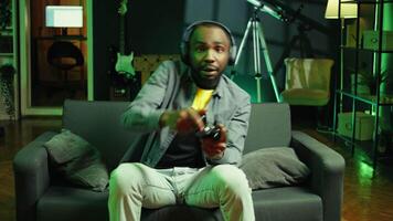 Enthusiastic gamer on couch using controller and headphones to play videogame in neon lit apartment. Player in RGB illuminated living room using high tech gaming console joystick, camera B video