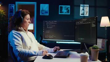 Woman listening music while doing software quality assurance, reading source code on computer screen. Developer wearing headphones enjoying podcasts and inspecting coding on desktop PC, camera B video