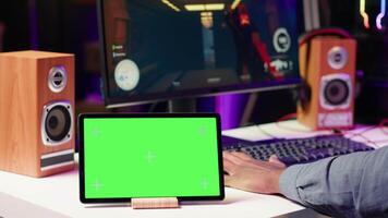 Chroma key tablet on desk next to man playing SF multiplayer videogame on computer with friends. Green screen device and gamer playing online with his Esports team in game video