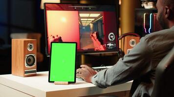 Green screen tablet next to professional streamer competing in online multiplayer videogame, streaming live at home. Chroma key device and man playing on PC, using headphones to talk with audience video