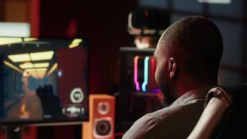 Man in neon lit apartment playing immersive games on gaming PC at computer desk, handheld camera shot. Gamer competing against enemies in internet multiplayer shooter, camera A video