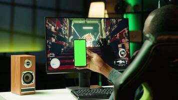 Man texting friends on green screen phone while playing first person shooter videogame at home. Gamer looking at mockup smartphone display, enjoying online discussion with mates video