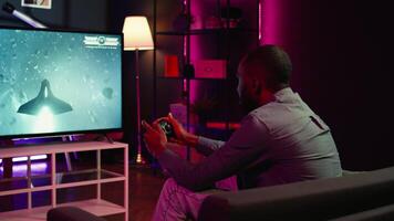 Gamer using controller and headphones to play first person shooter videogame on large smart TV. Man in home theatre using high tech gaming console gamepad to defeat foes, camera B video
