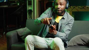 Enthusiastic gamer on couch using controller and headphones to play videogame in neon lit apartment. Player in RGB illuminated living room using high tech gaming console joystick, camera A video