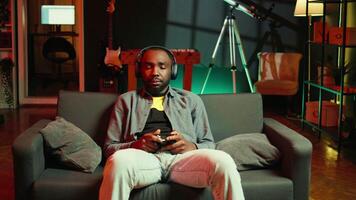 Tired gamer in RGB lit home late at night gaming with gamepad. Man sitting on sofa in neon illuminated apartment, playing videogames on console using controller, looking exhausted, camera B video